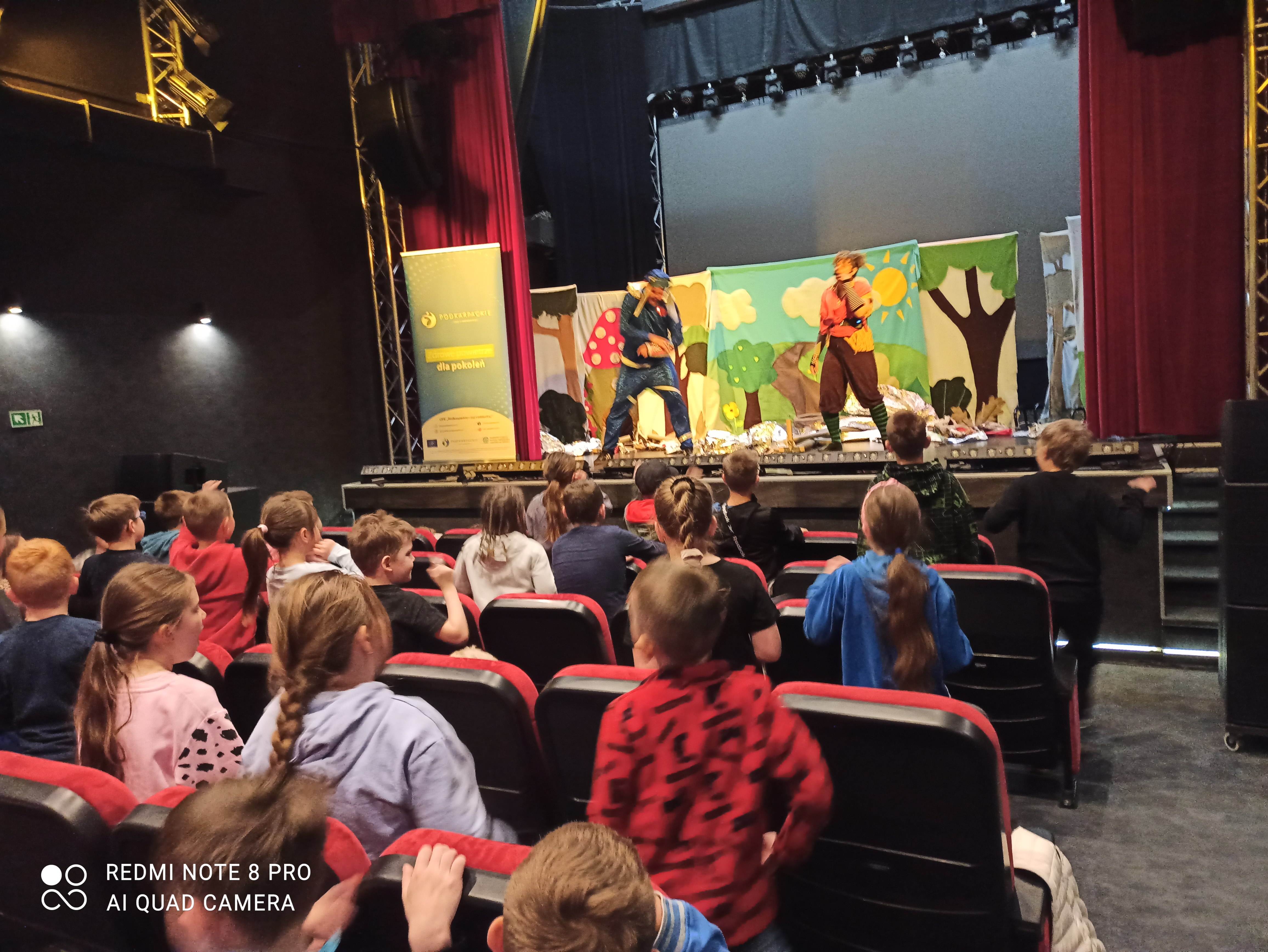 Grupa dzieci ogląda przedstawienie w teatrze. Na scenie dw&oacute;ch aktor&oacute;w w kostiumach. Scenografia z motywami przyrodniczymi. Widzowie siedzą na czerwonych fotelach.